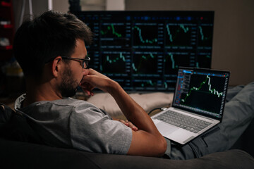 Thoughtful trader male in glasses focused on monitoring complex financial market data and cryptocurrency trends on laptop and large multi-screen setup, actively managing investments.