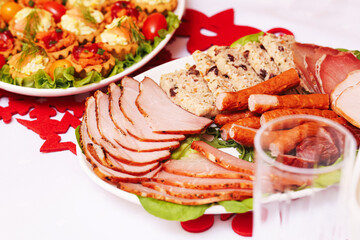 Cold cuts platter with roasted pork slices and sausage. Christmas table food with meat selection and salad in background. Holiday dinner spread with sliced ham and festive decoration.