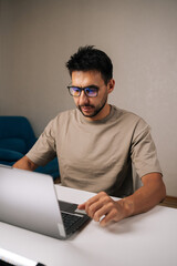 Bearded young businessman wearing glasses concentrating on work on laptop, typing on keyboard in remote home office setting during evening. Concept of remote workplace.