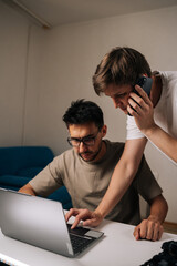 Vertical shot of two young entrepreneurs collaborating on startup project, discussing strategy and problem-solving while one man using laptop computer and other takes business call.