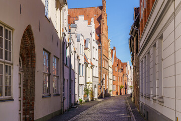 A narrow street in Lubeck Germany with a brick building on the left and a white building on the right. The street is lined with windows and has a brick walkway