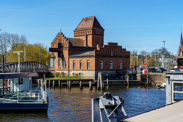 A large brick building with a red roof sits on a dock next to a boat in Lubeck Germany. The water is calm and the sky is clear