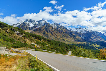 An asphalt road on the San Bernardino Pass in Switzerland The mountains are partially covered in snow. The mountains are also covered in a beautiful, colorful autumn blanket sky is blue and cloudy