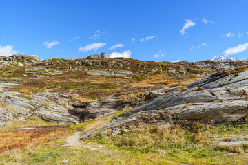 A rocky hillside with a small path leading up to it in Swiss Alps. The sky is clear and blue, and the grass is dry and brown. The scene is peaceful and serene, with the only sounds being the wind