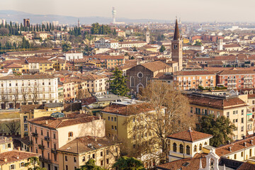A panoramic view of Verona, Italy, from above The Alpine mountains are visible in the background More visible are many square, multi-colored buildings and a red brick church
