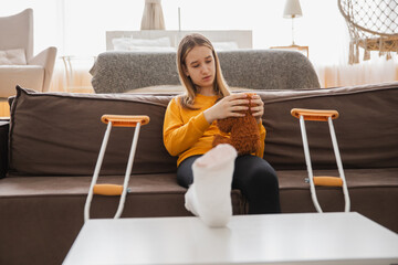 Young girl with broken leg cast sitting on sofa. Sad little kid hugging her teddy bear recovering from broken leg at home. Girl using cast and crutches. Accident injury fracture treatment foot bandage