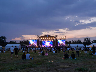 Obraz premium Wide-angle view of an outdoor music festival at dusk with a large illuminated stage, vibrant screens, and a crowd silhouetted against a purple-blue sunset
