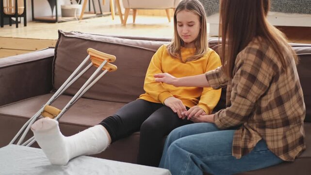 Girl with broken leg in cast sitting with mother on couch at home. Sad teenage kid recovering from broken leg. Girl using cast crutches for quick rehabilitation. Mom supporting daughter