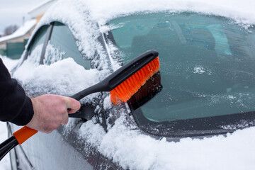 Hand holding a snow brush, clearing snow from a car window, showcasing winter weather conditions and the importance of vehicle maintenance during cold seasons