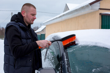 Male individual with short hair, wearing a black vest, is clearing snow from a car windshield using a brush, showcasing winter maintenance and vehicle care in a snowy environment