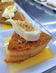 A piece of traditional Turkish bread pudding or ekmek kadayifi topped with a thick layer of clotted cream served on a white plate with sweet syrup in a restaurant display setting