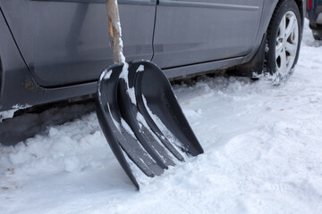 Black snow shovel resting against a gray vehicle, surrounded by fresh snow, showcasing winter weather challenges and the need for snow removal tools in cold climates © Krystsina
