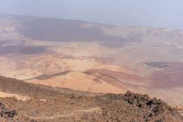 Aerial view of Teide volcano caldera landscape shows reddish volcanic hills, sandy dunes, rocky ridges, and distant haze under clear blue sky from lookout point in Tenerife, Canary Islands, Spain.