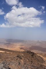 Vista from lookout point reveals Teide volcano caldera with reddish volcanic slopes, sandy expanses, rocky ridges, distant haze, scattered clouds, and blue sky in Tenerife, Canary Islands, Spain.