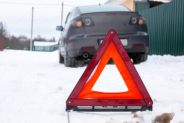 Warning triangle placed in snow on roadside, with a car parked in the background, indicating vehicle breakdown and the need for caution in winter conditions