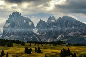 Dramatic mountain landscape with towering rocky peaks, green alpine meadow and dense forest under a cloudy sky. Scenic nature background featuring majestic mountains, rolling hills.