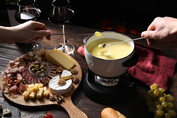 Couple dipping piece of bread and grape into fondue pot with delicious melted cheese at wooden...