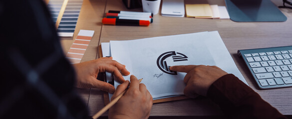A collaborative workspace shows hands sketching a logo design with color palettes and digital tools in a modern office, emphasizing creativity and teamwork in branding. SACTR