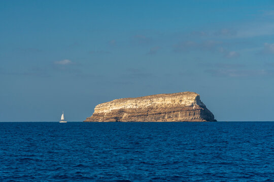 Isolated rocky island in deep blue sea with sailboat on horizon - Powered by Adobe