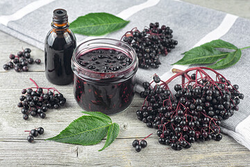 Delicious homemade black elderberry syrup in glass jar and bunches of black elderberry with green leaves on wooden desk.