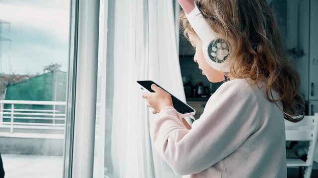 Little girl with headphones using smartphone by window at home