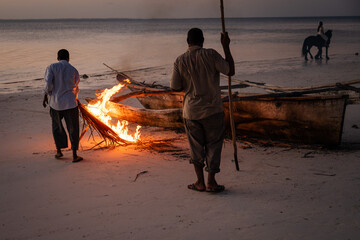 Two local men use a small fire on a sandy beach at twilight to repair or seal a traditional wooden outrigger boat in Zanzibar