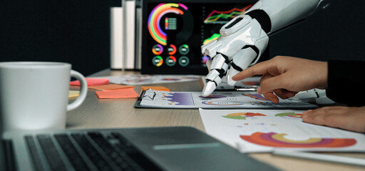 A futuristic scene showing a robotic hand aiding a human in analyzing colorful graphs and charts on a sleek desk, highlighting the cooperation between technology and creativity.Noogenesis