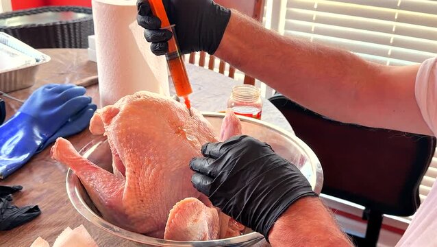 Close-up video of a raw whole turkey being injected with marinade using a syringe during food preparation for a holiday meal. Hands wearing protective gloves are shown seasoning poultry in a kitchen.