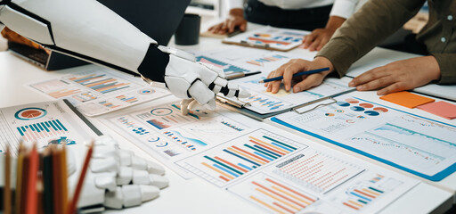 A robotic arm aids human colleagues in a modern office environment, focusing on data analysis and strategic planning with various reports and charts displayed.Noogenesis