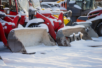 Snowy construction equipment yard with red tractor loaders and metal buckets, close up of heavy machinery attachments and tires stored outdoors in winter