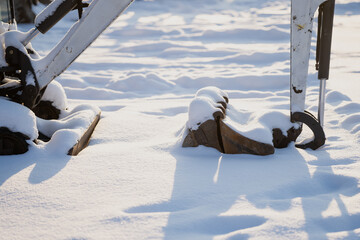 Snow covered machine attachments resting on ground, close up of metal parts and hydraulic arm pieces casting long shadows on fresh snow in winter sunlight