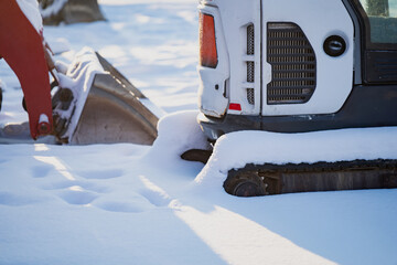 Snow covered tracked skid steer loader parked in deep snow, close view of rubber track and rear body panel with excavator bucket blurred in winter yard