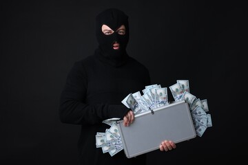 Masked thief with briefcase full of money on black background