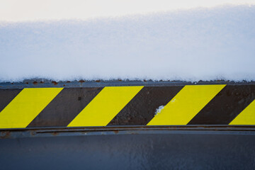 Snow covered steel edge with diagonal hazard stripe pattern in yellow and black, close view of industrial safety sign marking on equipment in winter light