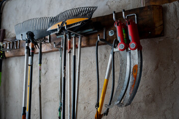 Wall mounted rack holding rakes, long handle yard tools and red handled sickle saws in garden shed, organized equipment hanging from wooden board on plaster wall