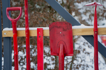 Red metal snow shovels and ice tools hanging on wooden rack outdoors with light snow showing winter maintenance equipment storage and seasonal work tools