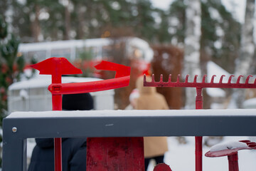 Red metal outdoor tool rack with snow covered rakes and brackets, close up of equipment frame with blurred people walking in snowy yard background