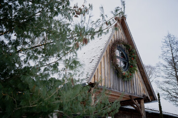Timber house facade with round window and festive wreath, winter roof with snow and soft overcast sky, evergreen foliage partly covering view