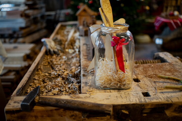 Rustic workshop still life showing clear jar gift with ribbons and wooden ornament, sitting on old workbench with wood shavings, hammer and craft clutter