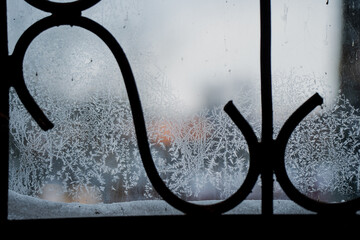 Frost patterns on window glass with black wrought iron railing silhouette, cold winter texture and blurred outdoor background seen through icy pane