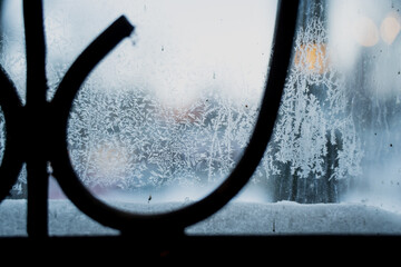 Icy window pane with delicate frost patterns and snow buildup on ledge, shallow depth of field showing cold winter atmosphere with blurred scene outside