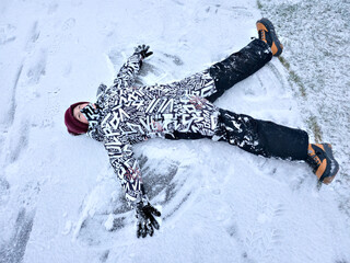 A happy child lies in the snow with snow angel wings. An excited boy in winter clothes lies in the snow.