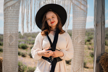 Woman stands in a rural setting wearing a hat and posing near decorative fabric in sunlight