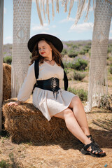 Young woman stands in outdoor setting wearing hat and dress while holding a bow of fabric, surrounded by natural scenery in sunny weather
