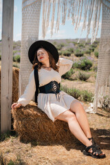 Young woman stands in outdoor setting wearing hat and dress while holding a bow of fabric, surrounded by natural scenery in sunny weather