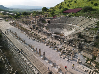 Aerial view presents the Odeon of Ephesus in Turkey Stone seating curves around the orchestra People explore the stage and grand facade Columns and blocks fill the forecourt Historic stones line the © MikeDrone