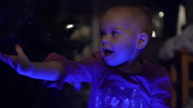 blue-eyed girl rejoices at the bright blue lights of the garland on her lap, joyfully raises her hands, looks at the camera and smiles, opens her mouth and laughs cheerfully