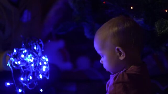 A baby girl looks at the bright blue New Year s garland on the floor for the first time. enthusiastically and incomprehensibly opens his mouth, looks into the camera, touches the light bulbs and