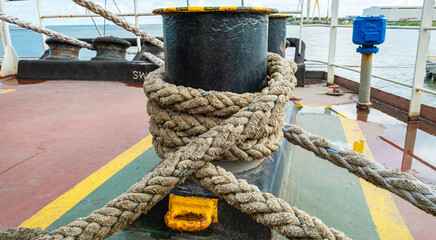 Mooring rope wrapped around a black bollard on a ship deck. Marine industrial detail with textured nautical cable, yellow safety markings, and harbor background