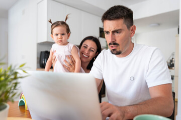 Father working anxiously from home with a laptop, multitask parenting while mother holds concerned toddler looking at a screen in a modern bright kitchen office setting